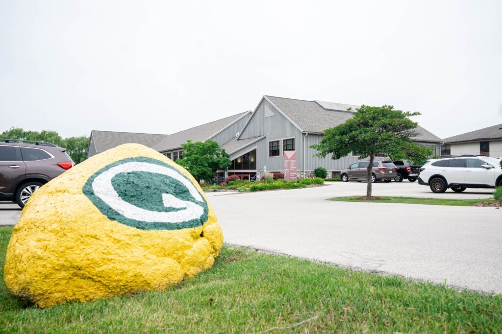 Kittleson Orthodontics office exterior in Mequon, featuring a large yellow rock with a green Packers logo, surrounded by greenery and parked cars.