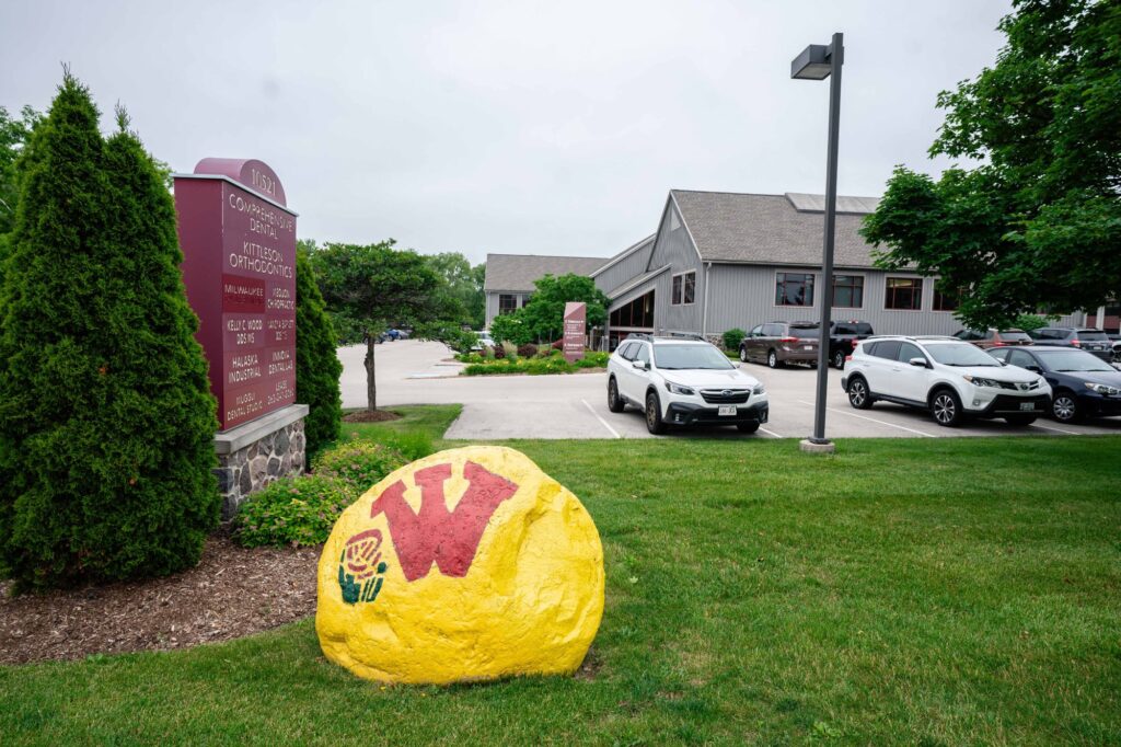 Kittleson Orthodontics office exterior in Mequon featuring a colorful yellow rock with a "W" logo, surrounded by green landscaping and parked vehicles, highlighting the modern orthodontic facility.