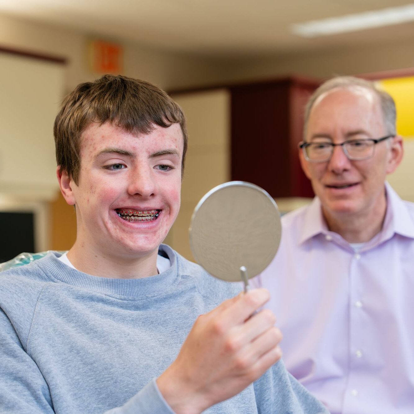Teenager with braces smiling while looking in a mirror, with Dr. Kittleson observing, in a Mequon orthodontics office setting.